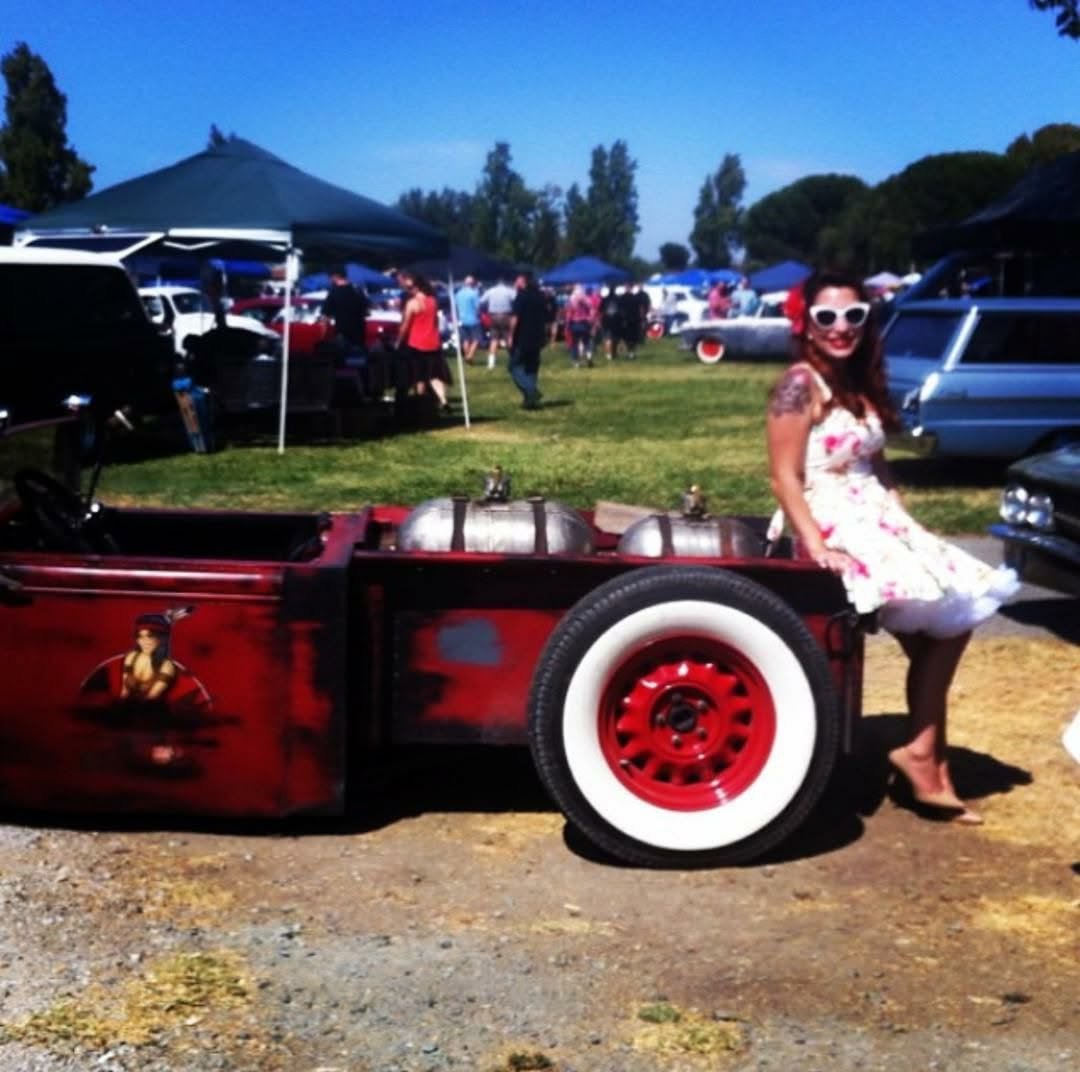 Jennifer in a floral vintage dress and cat eye sunglasses, leaning against a rat rod at a car show.