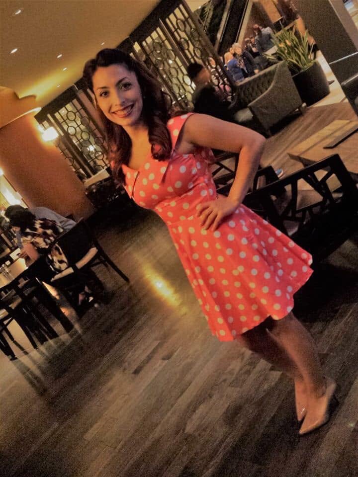 Jennifer in a coral polka dot dress, one hand on her hip, smiling.